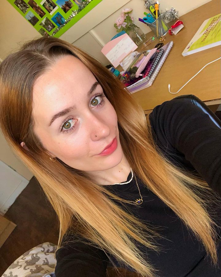 University Student, Ella smiling and looking into the camera while taking a selfie. She has brown hair, a black top and a gold necklace. The beginning of her day.
