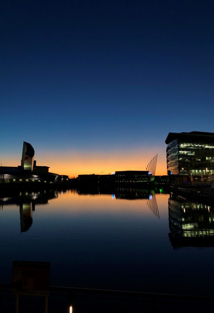 Nighttime view of Salford Quays Media City University with illuminated buildings and reflection on the water in the foreground. This is what University students see at the end of the day